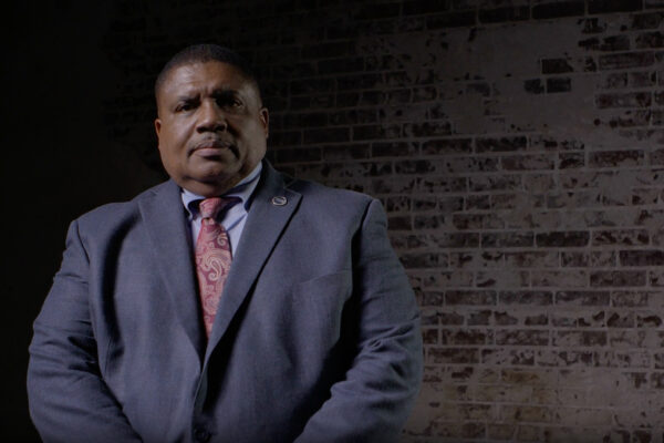 Portrait of a man in a blue suit and red paisley tie standing against a dark brick wall, looking at the camera.